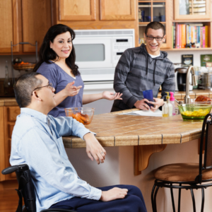 A man in a wheelchair, a brunette woman standing, and a younger man with glasses talk and laugh in a kitchen